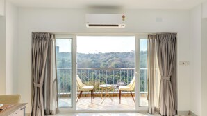 Una habitación amplia con puerta de vidrio corredera que da a un balcón con mesa y sillas, un equipo de aire acondicionado y vistas a un paisaje montañoso.