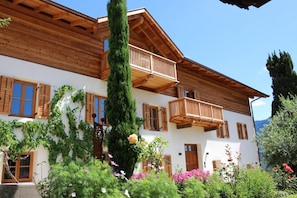 A two-story wooden building with balconies, a garden with flowers, and a clear sky. - Apartment 'Greiterhof Weingarten' with Mountain View and Wi-Fi (Lana)