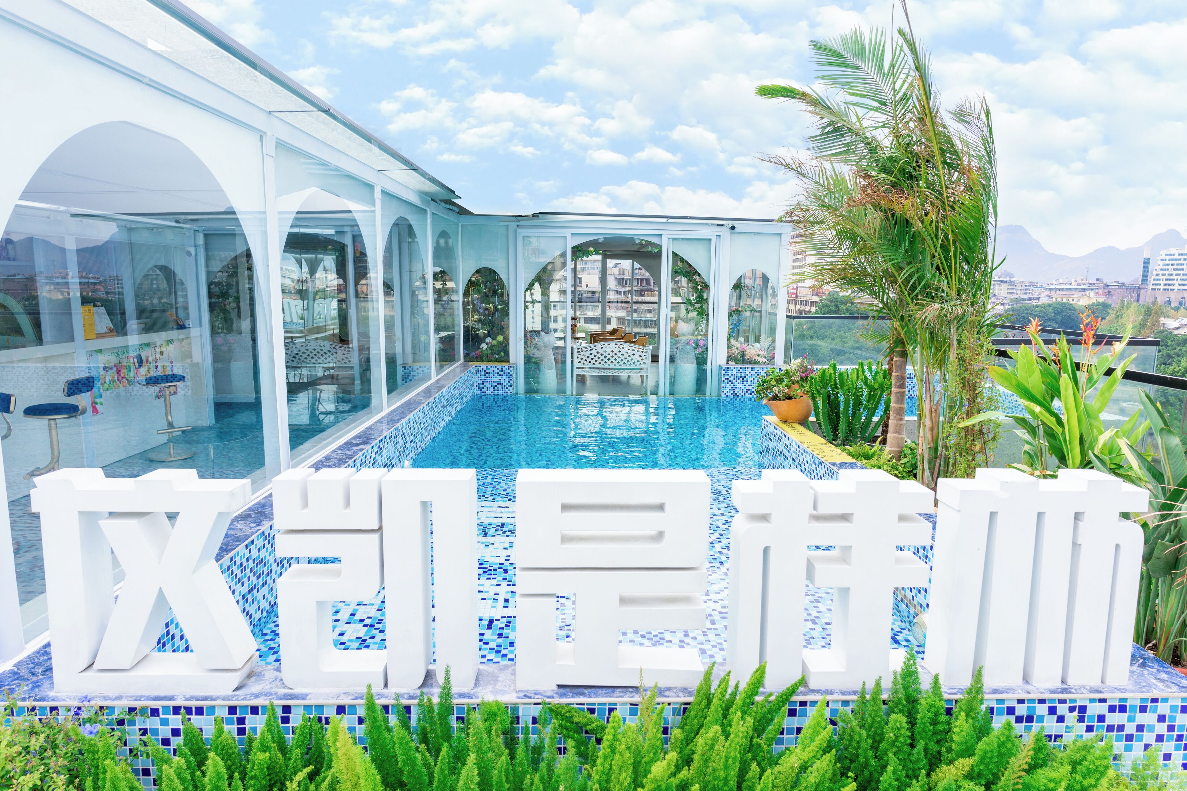 A modern hotel with a large glass-enclosed pool, outdoor seating, and tropical plants.