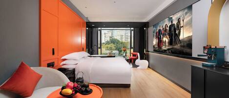 A modern hotel room with a large bed, a dining table with fruit, a coffee machine, and a city view.