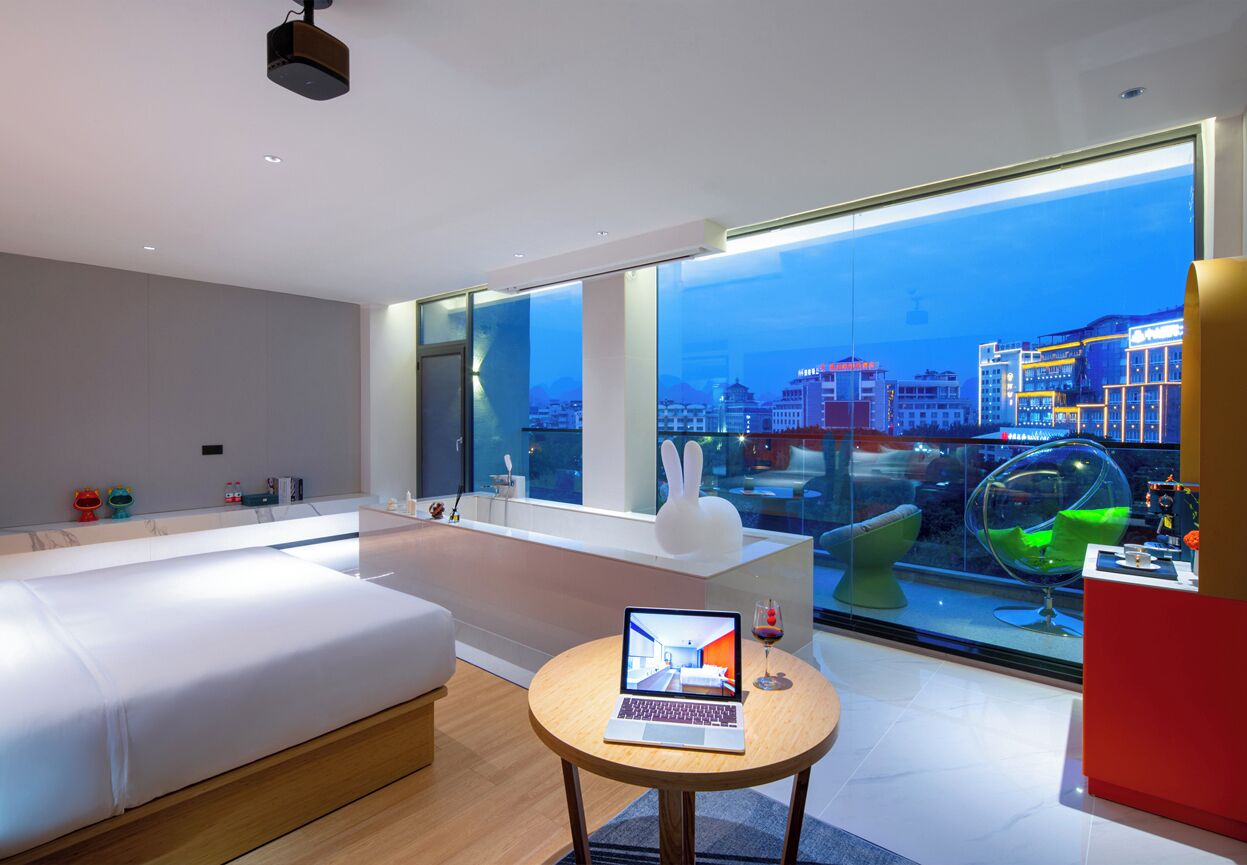 A modern hotel room with a large window, a laptop on a small table, and a view of the cityscape.