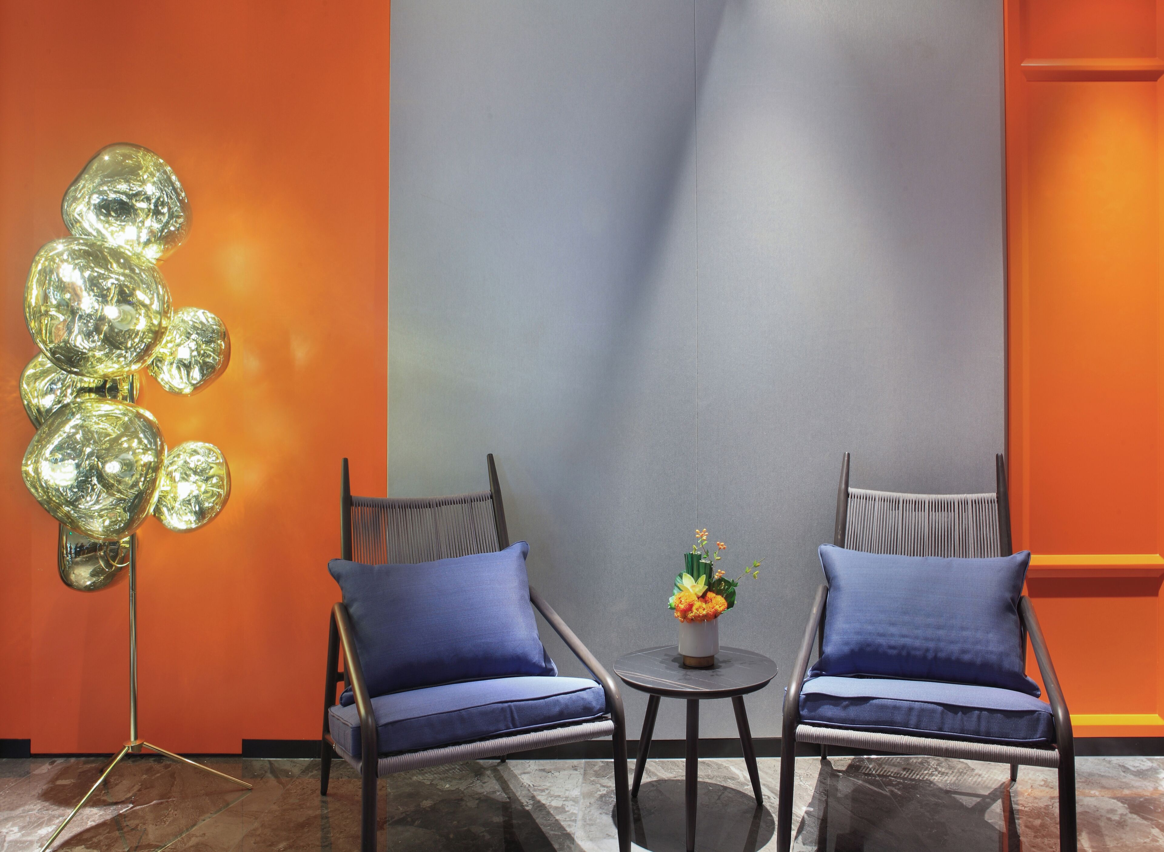 Una habitación con dos sillones azules, una pequeña mesa redonda con un florero y una lámpara moderna frente a una pared bicoloreada.