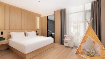 A modern hotel room with a wooden bed, a small bedside table, a nightstand, a wardrobe, a window with curtains, and a children's play tent.