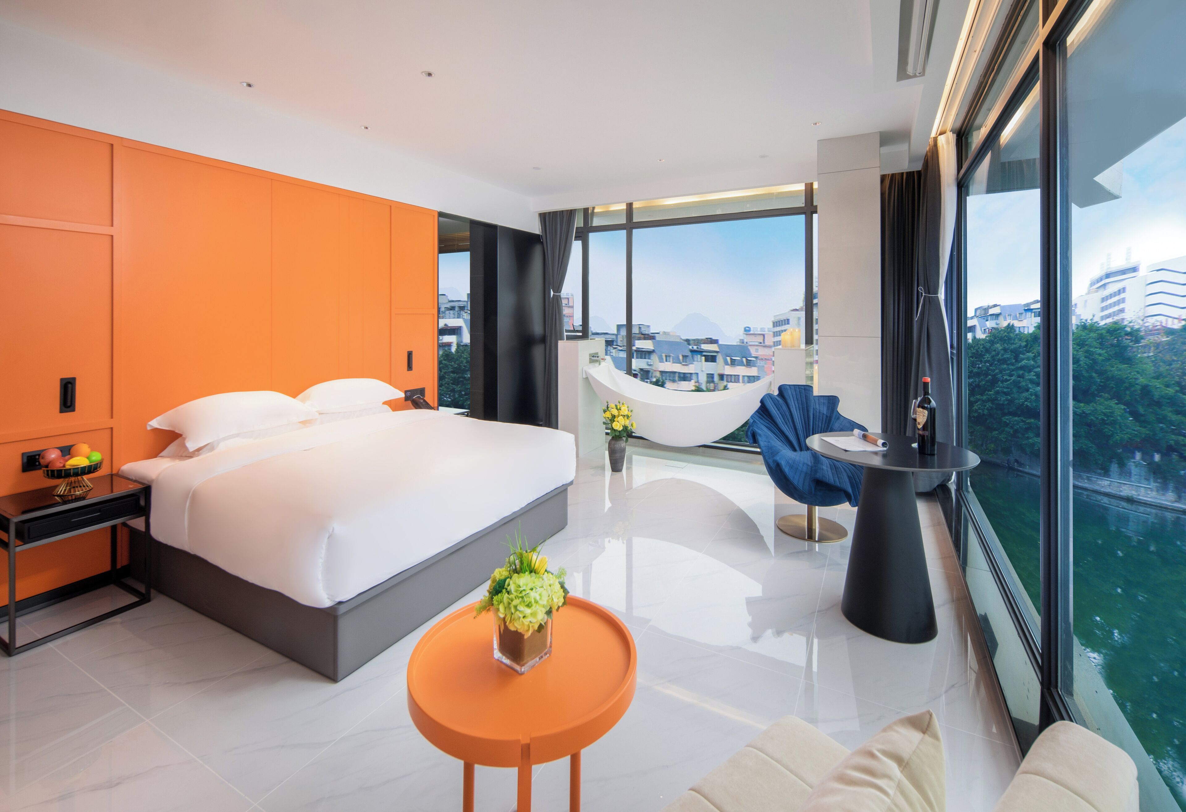 A modern hotel room with a large bed, a small orange table, a sofa, and a view of the city through large windows.