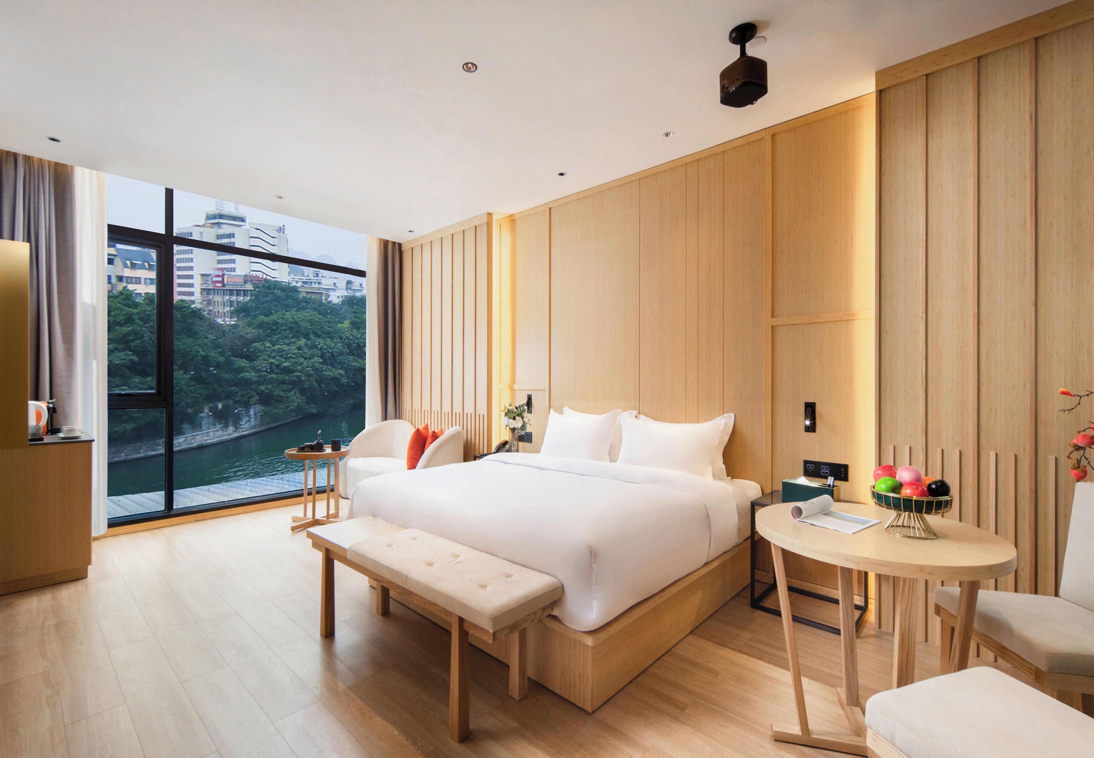 Una habitación de hotel moderna con una cama grande, un banco, una mesita y vistas a un paisaje urbano a través de amplios ventanales.