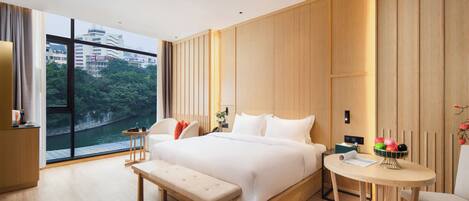 Una habitación de hotel moderna con una cama grande, un banco, una mesita y vistas a un paisaje urbano a través de amplios ventanales.