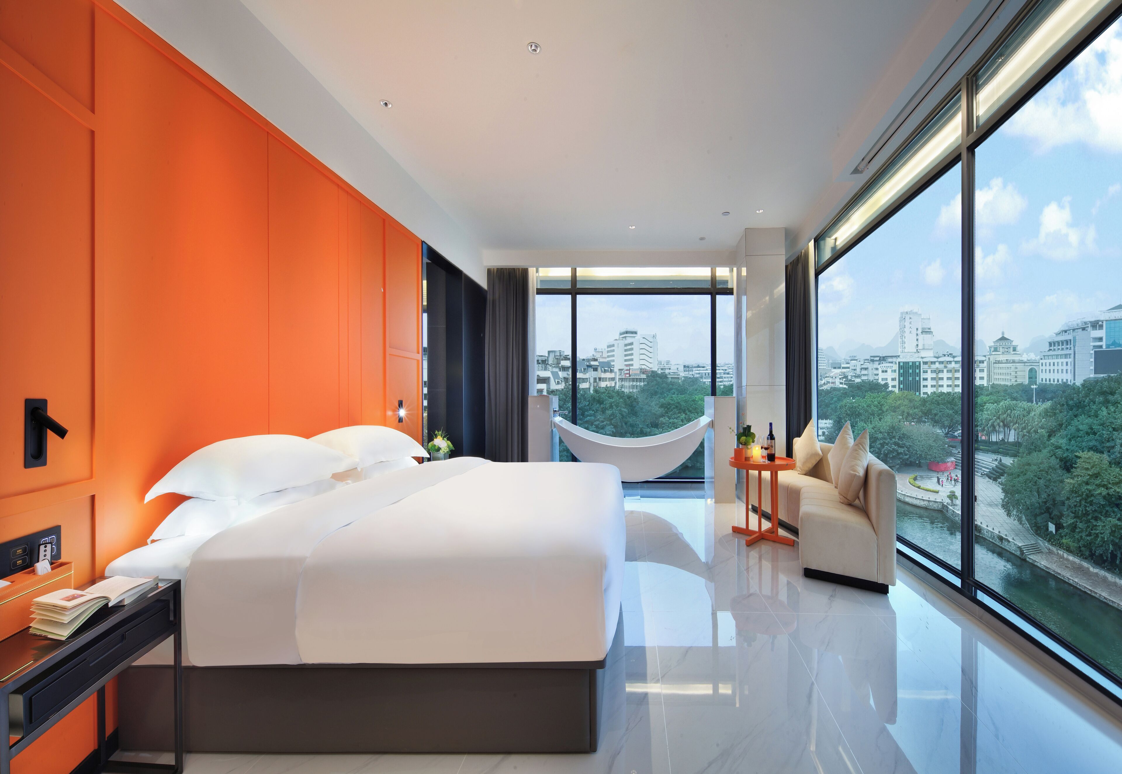 A modern hotel room with a large bed, a desk, and a view of the city through large windows.
