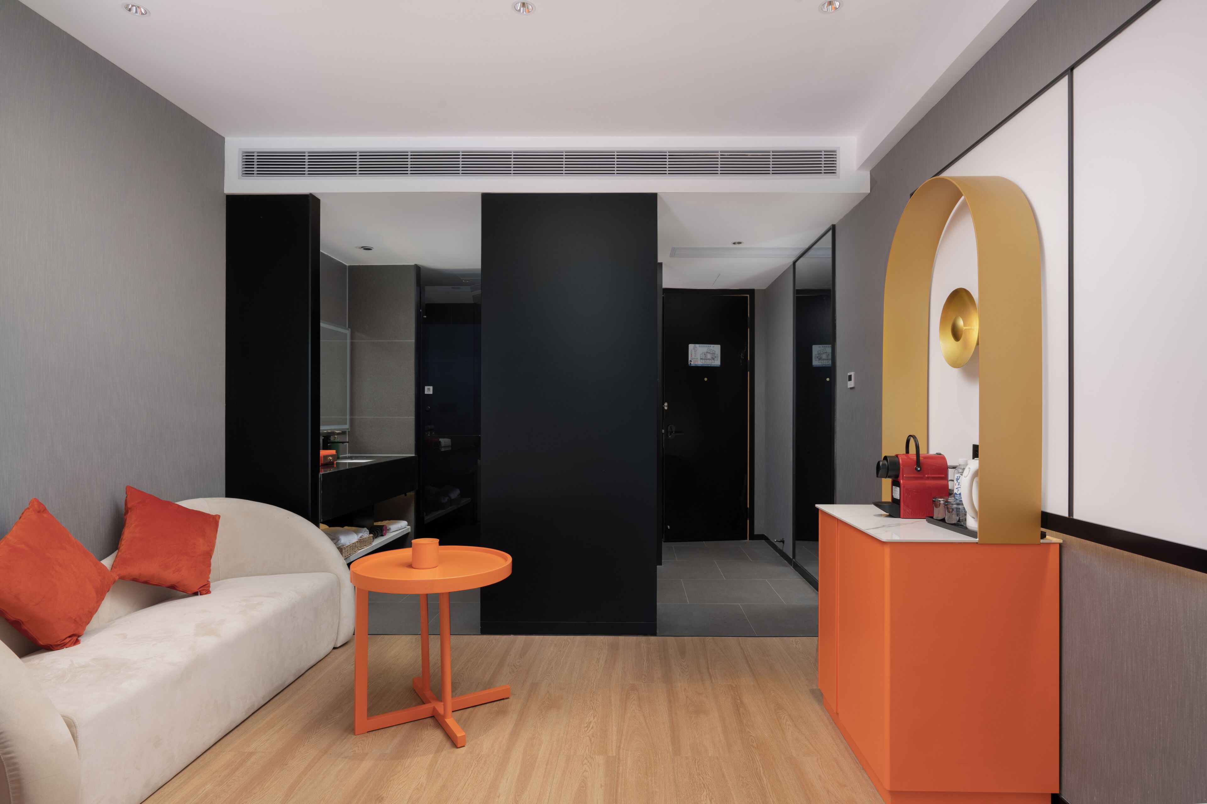 Une chambre d’hôtel moderne avec un canapé, une petite table orange, un comptoir de réception et un grand miroir.