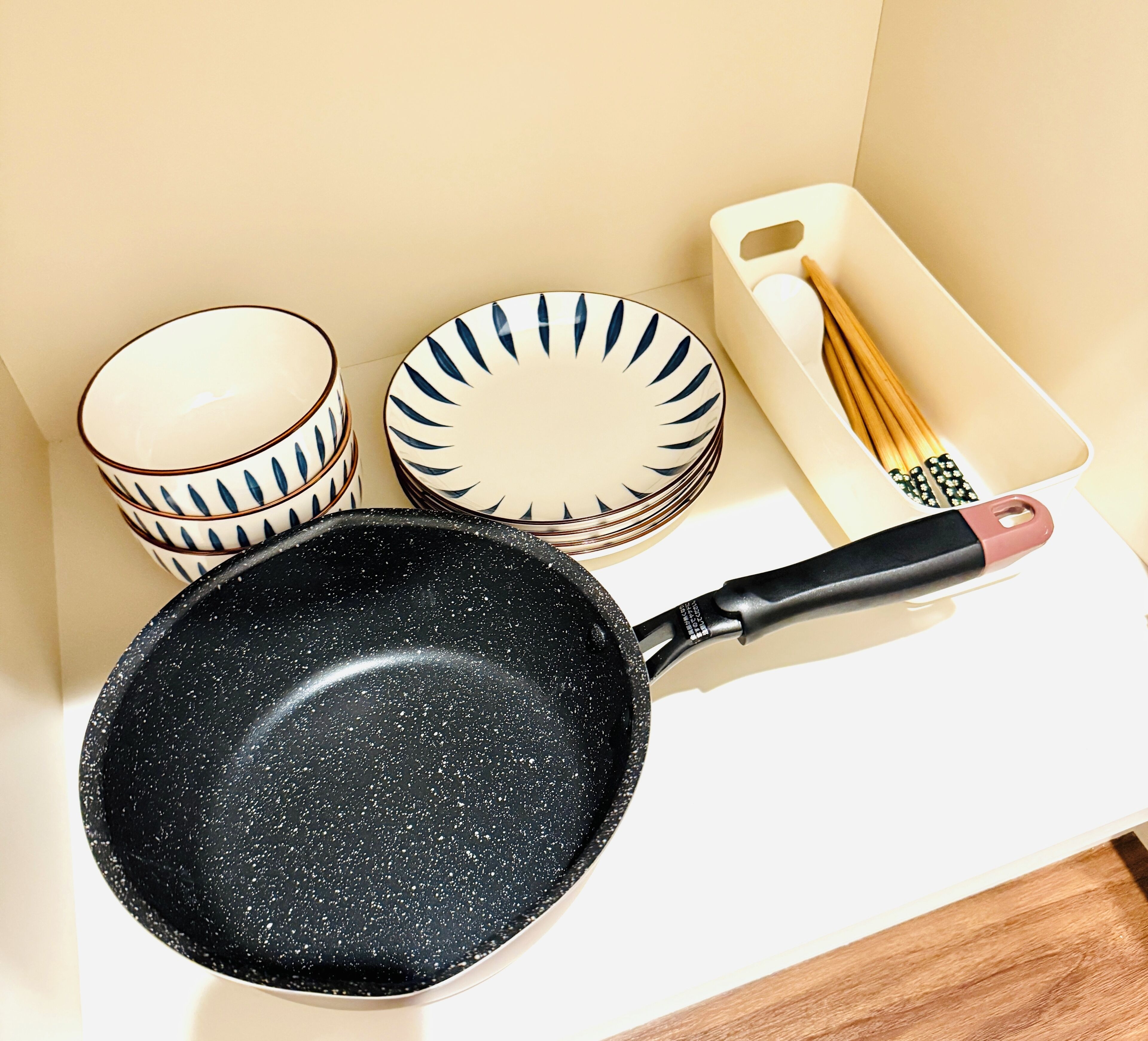 Una encimera de cocina con una sartén negra, platos blancos y azules apilados, y una tabla de cortar de madera con palillos.