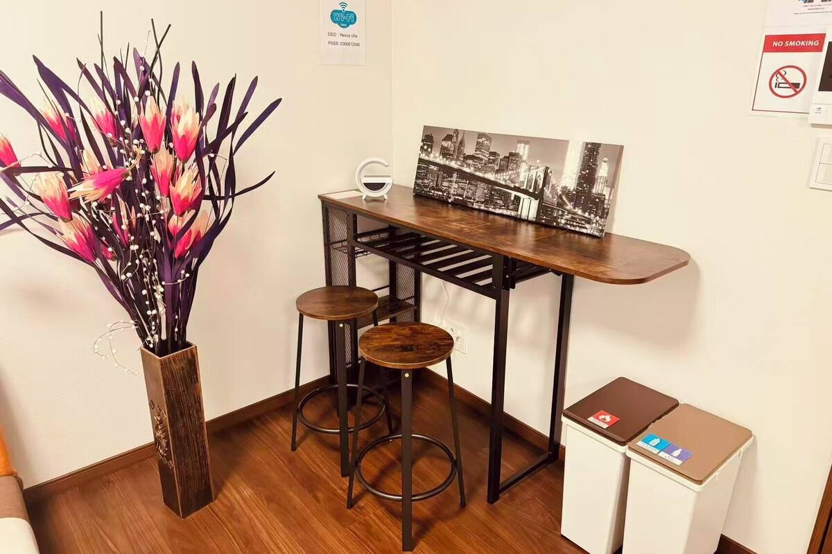 A small office area with a desk, two stools, a vase with flowers, and a cityscape painting.