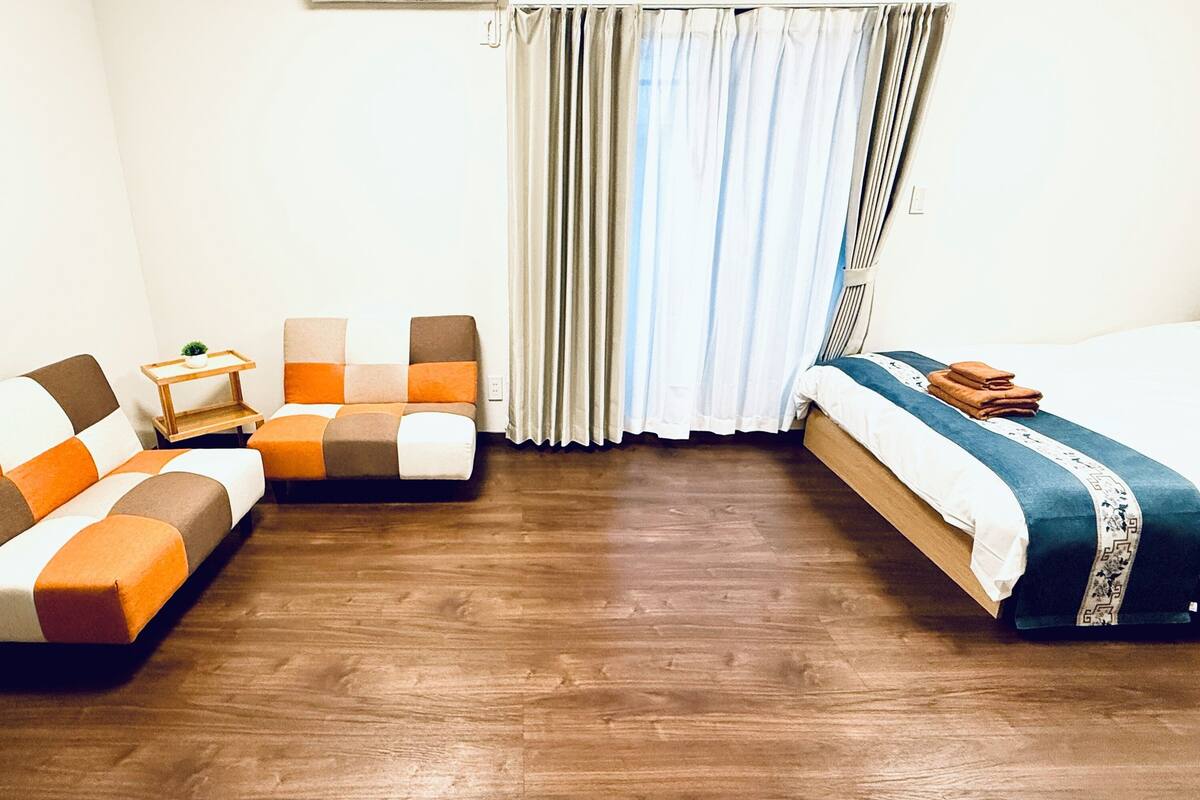 A hotel room with a wooden floor, a sofa with orange and white cushions, a small wooden side table, a bed with a blue and white bedspread, and a window with curtains.