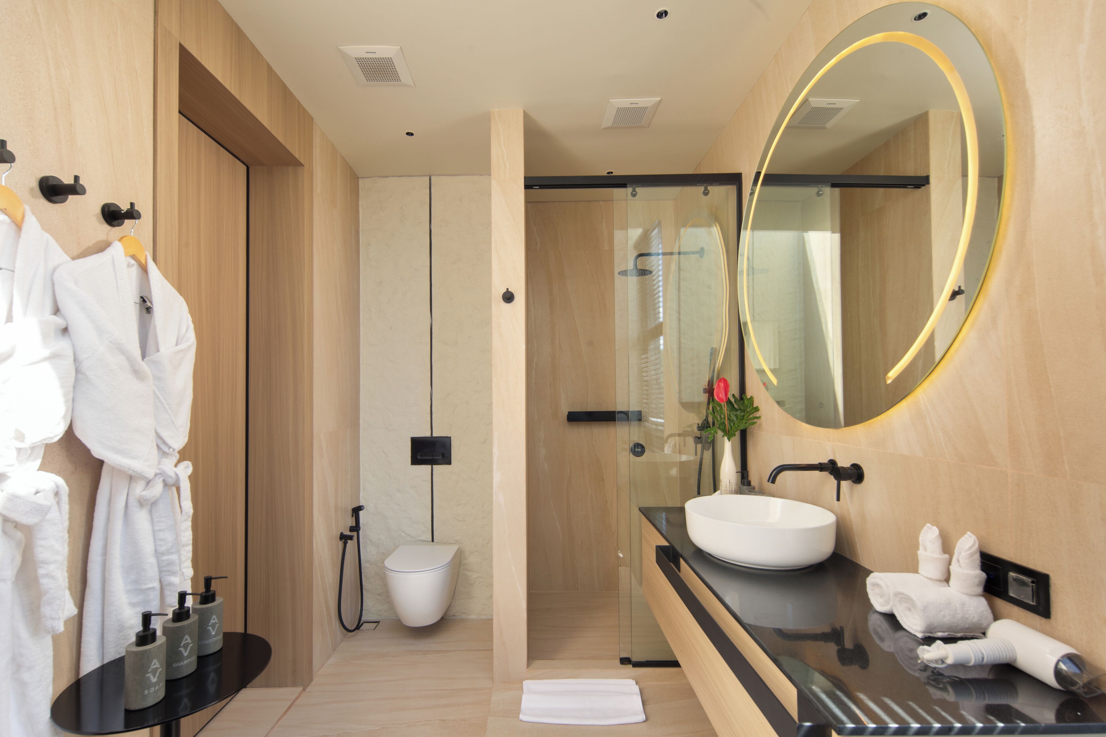 Un baño moderno con ducha envuelta en vidrio, un espejo redondo con marco dorado, un lavabo blanco y una encimera negra.