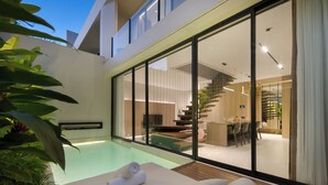 Un área de piscina moderna con una silla de descanso, una toalla y un comedor visible a través de grandes puertas de vidrio.