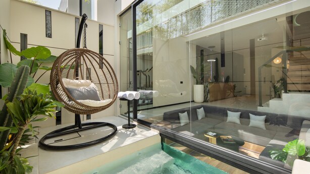 Un espacio exterior moderno con una silla colgante, una piscina con cubierta de vidrio y una zona de estar acogedora con cojines.