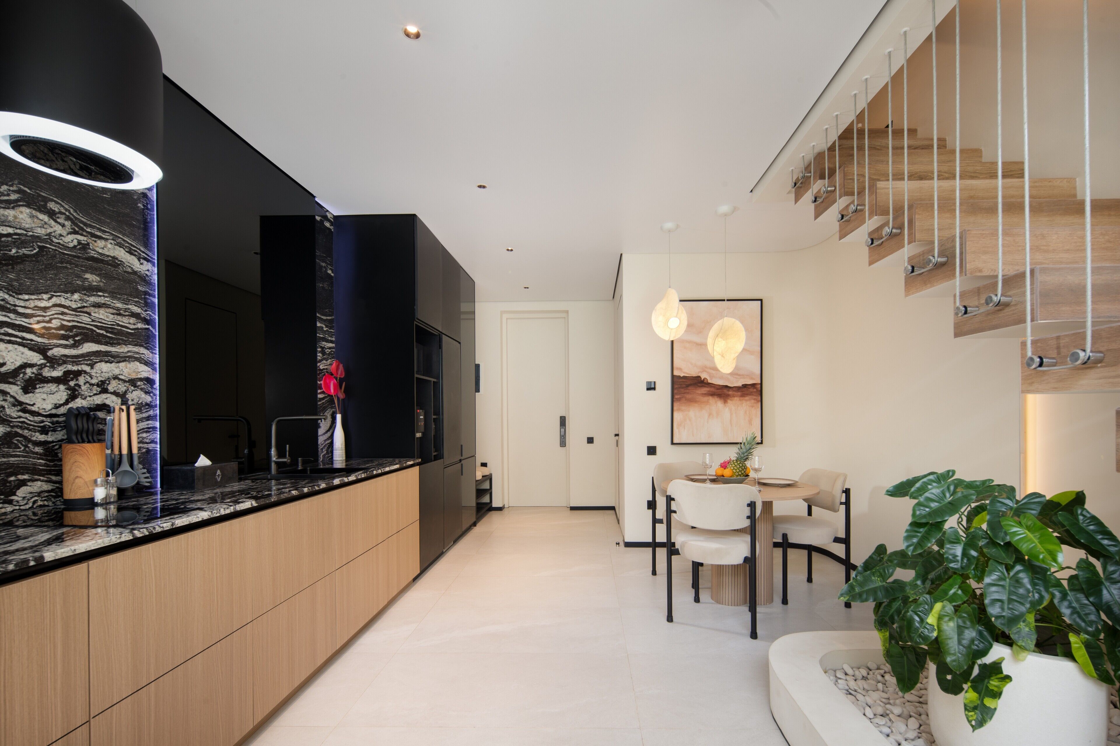 A modern kitchen with a marble countertop, wooden cabinets, and a built-in oven. There is a dining area with a white table and chairs, and a large potted plant.