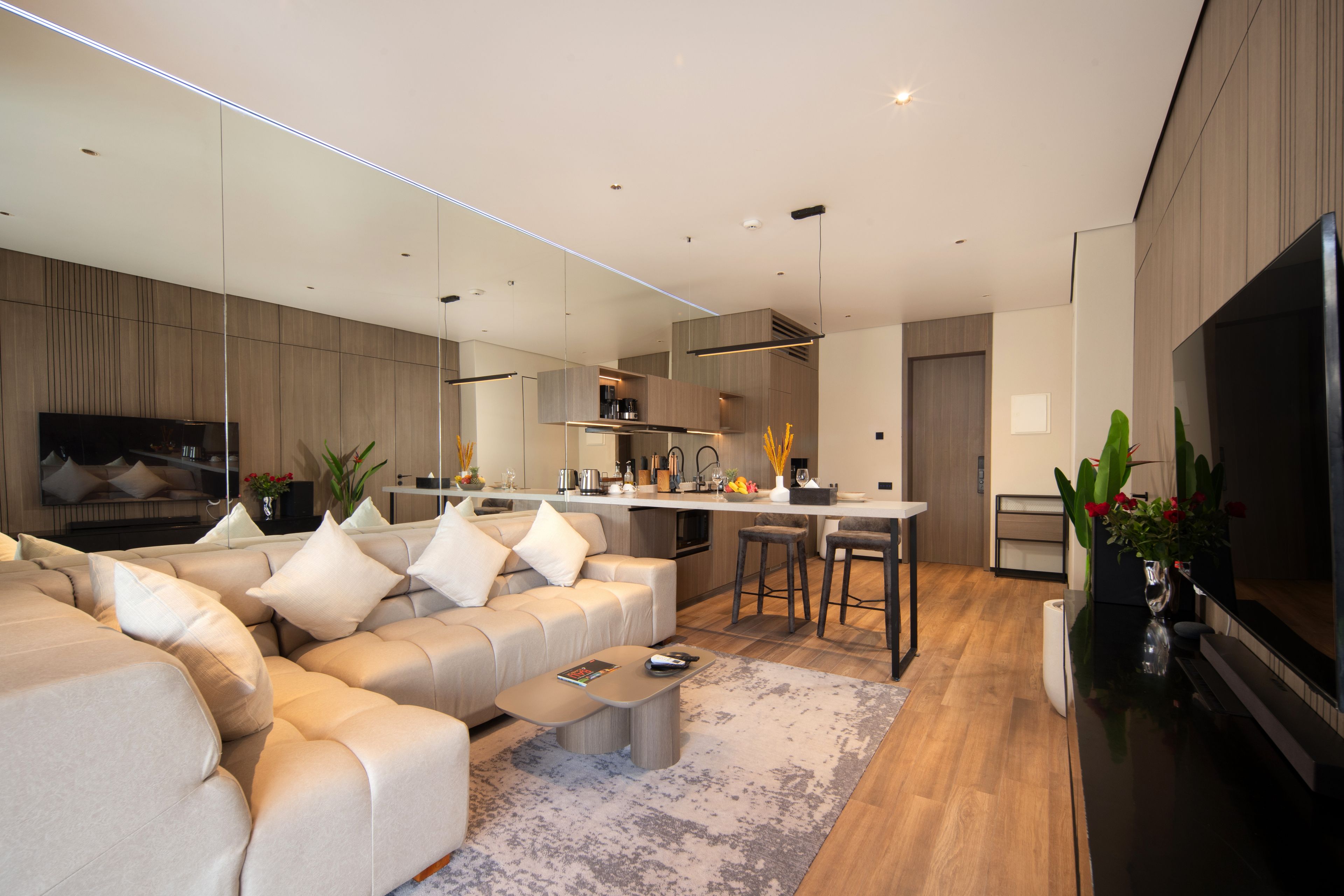 A modern living room with a large flat-screen TV, a beige sofa, and a kitchen area with bar stools.