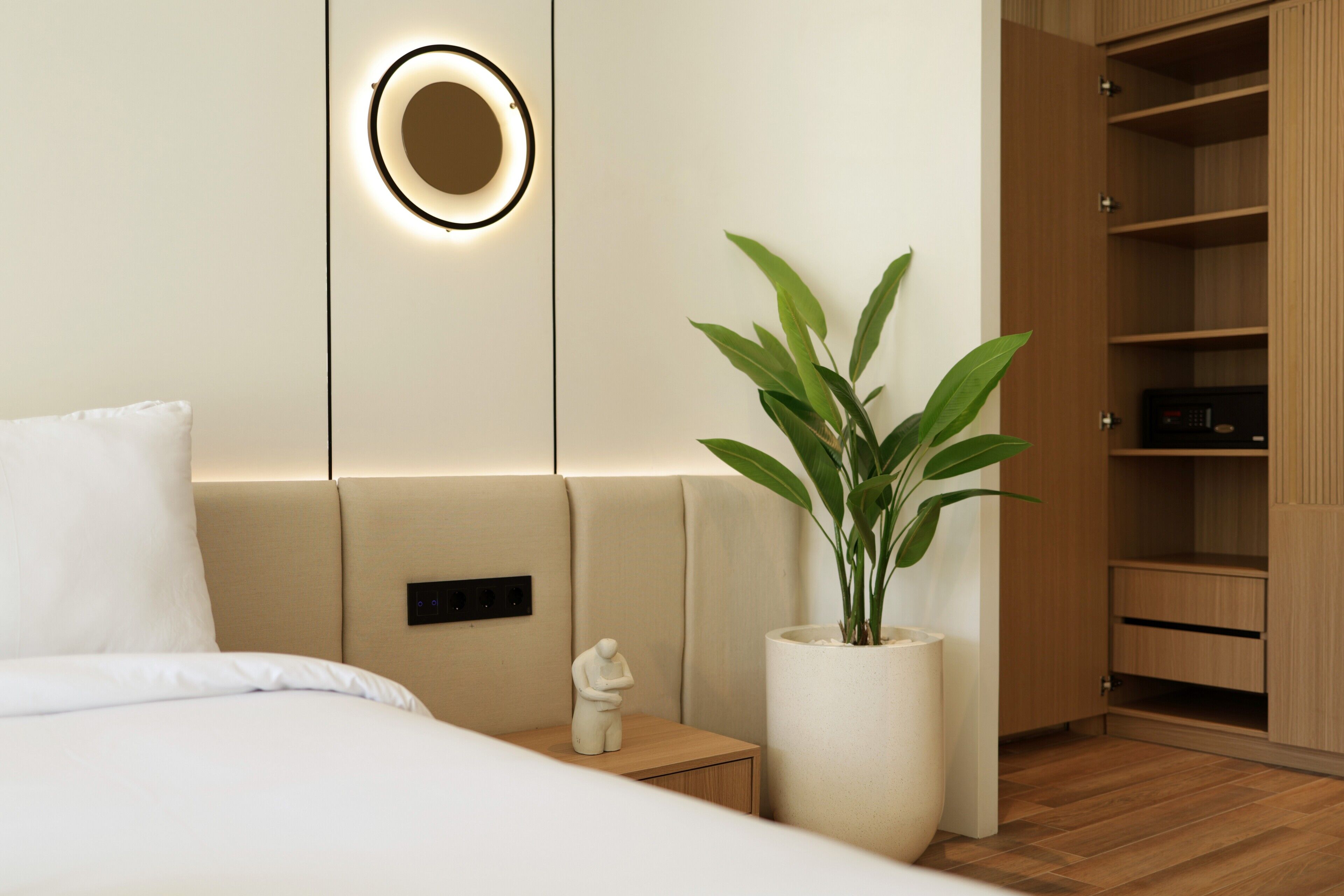 Une chambre d’hôtel moderne, équipée d’un lit, d’une table de chevet, d’une grande plante en pot, d’un luminaire fixé au mur et d’une armoire en bois.