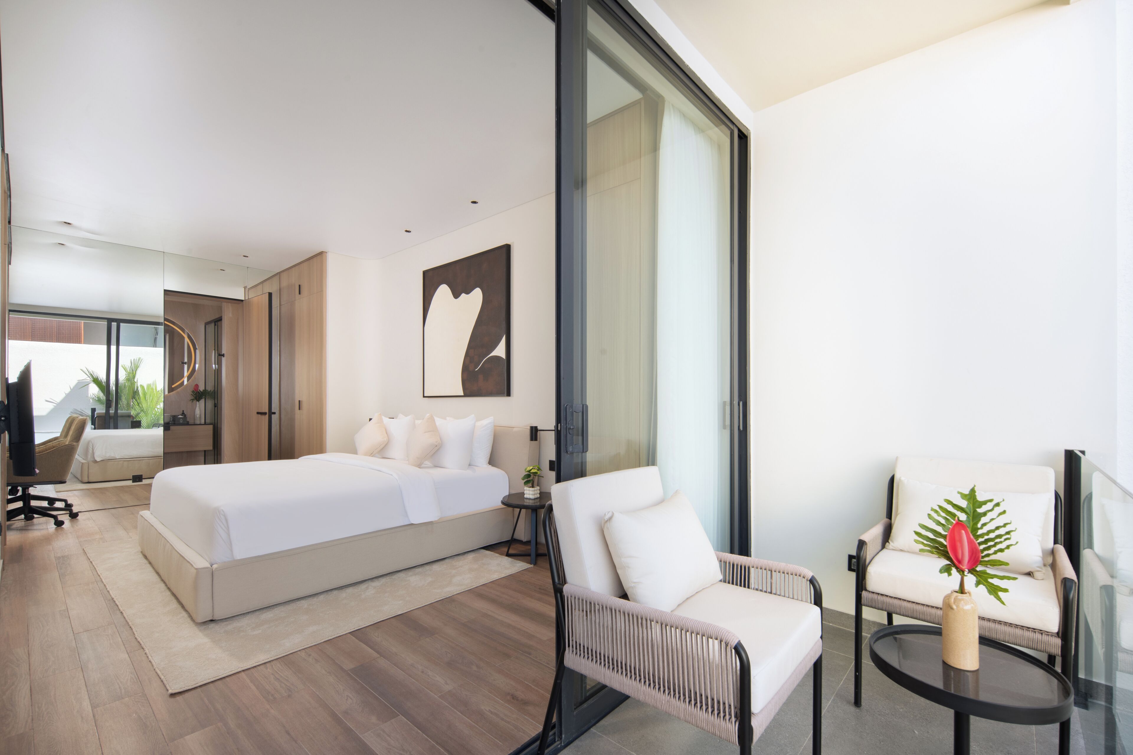 A modern hotel room with a large bed, two chairs, a small table, and a large window.
