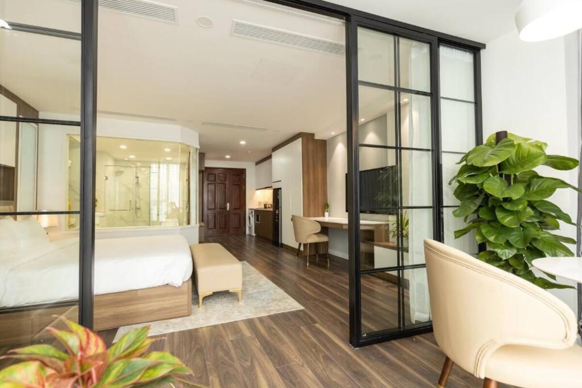 Una habitación de hotel moderna con una cama, un escritorio y una silla. Hay un ventanal grande y una planta en la esquina.