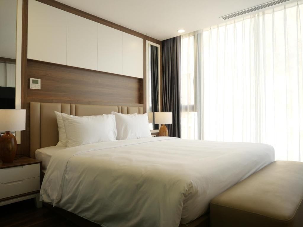 Une chambre d’hôtel avec un grand lit, deux tables de chevet avec des lampes, une tête de lit en bois et une fenêtre avec des rideaux légers.