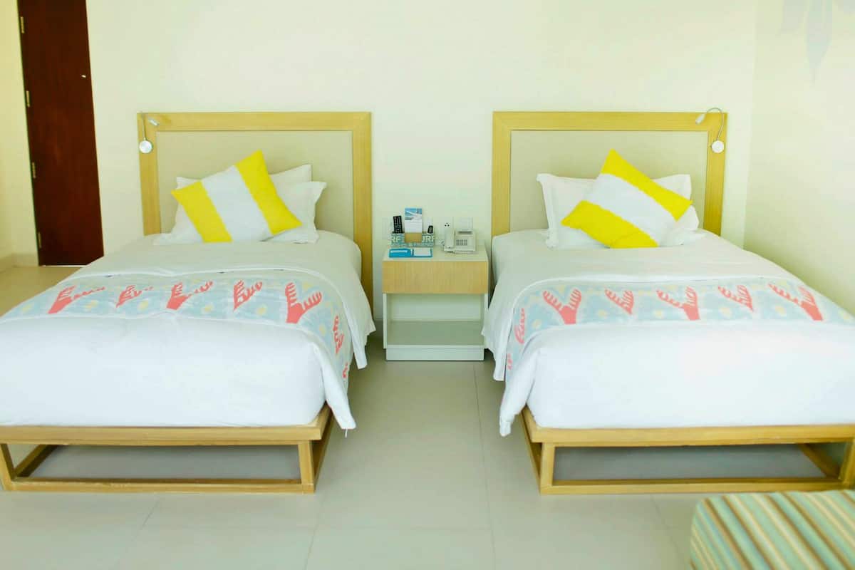 Habitación de hotel con dos camas, cada una con cabecero de madera, ropa de cama blanca y almohada amarilla y blanca. Hay una mesita de noche con un teléfono y una lámpara pequeña.