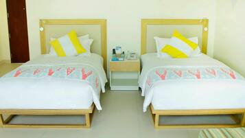 Habitación de hotel con dos camas, cada una con cabecero de madera, ropa de cama blanca y almohada amarilla y blanca. Hay una mesita de noche con un teléfono y una lámpara pequeña.