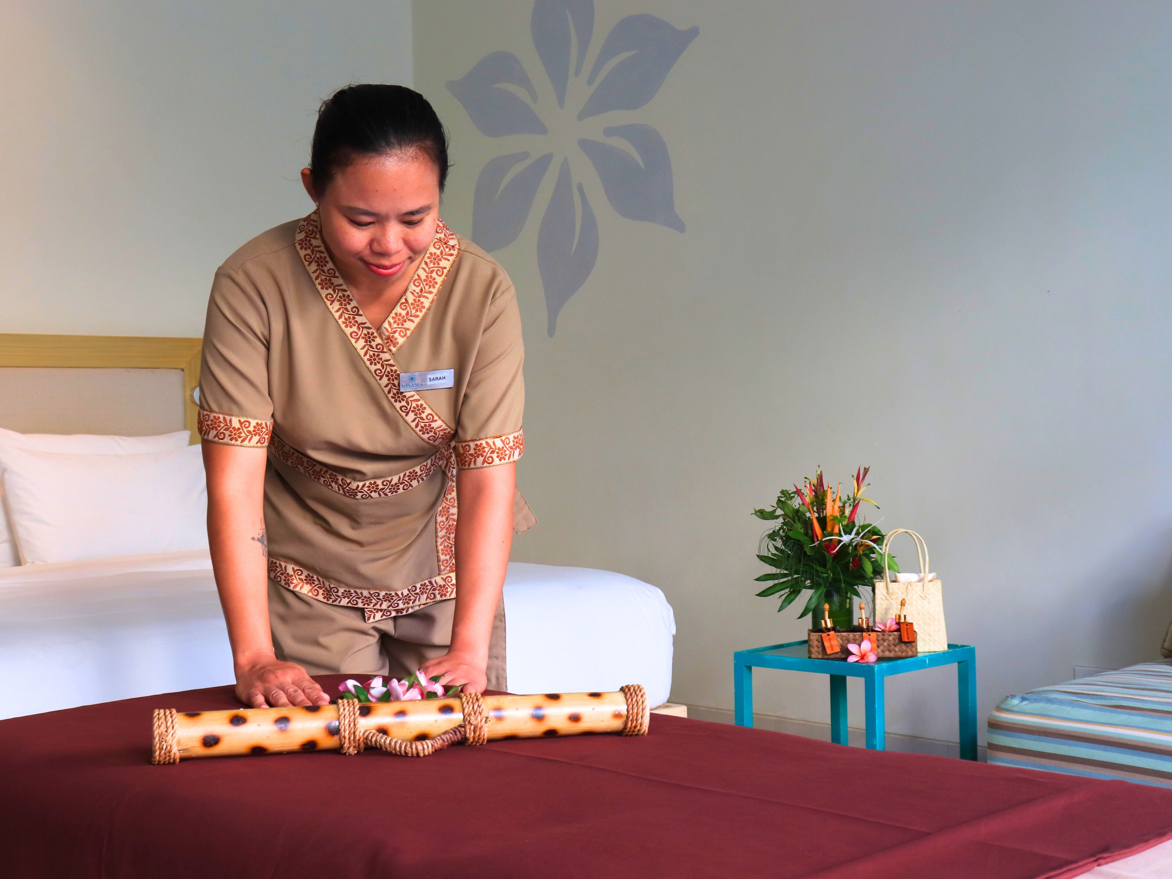 A person in a room with a bed, a small table with a plant and a bag, and a wall with a floral design.
