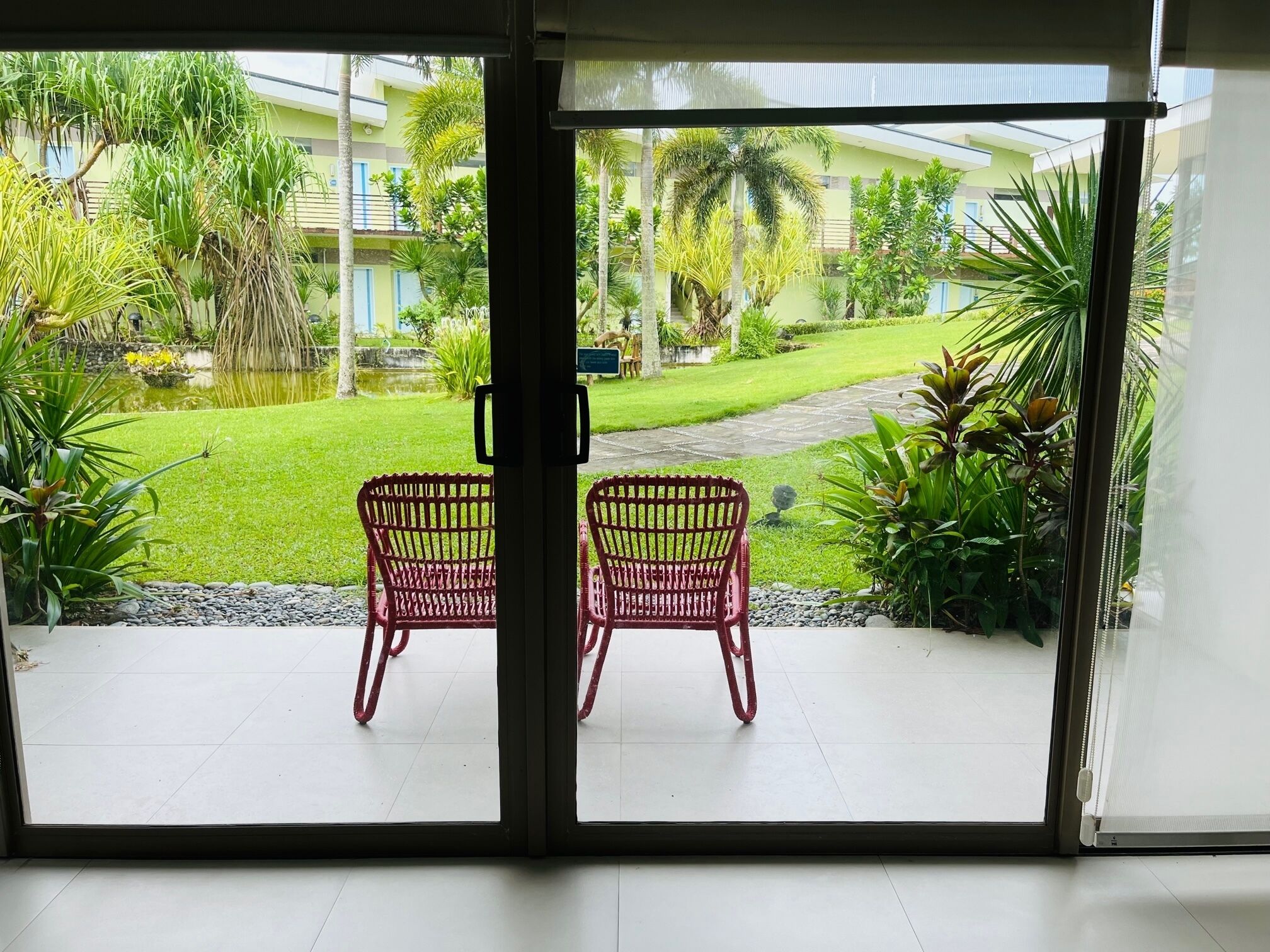 Vista attraverso una porta vetrata su un patio con due poltrone in vimini, uno stagno e un giardino curato, ricco di piante e prato.