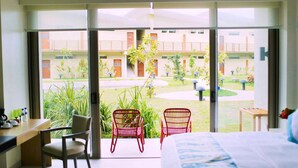 A hotel room with a bed, desk, two red chairs, and a view of a courtyard with plants and a building in the background.