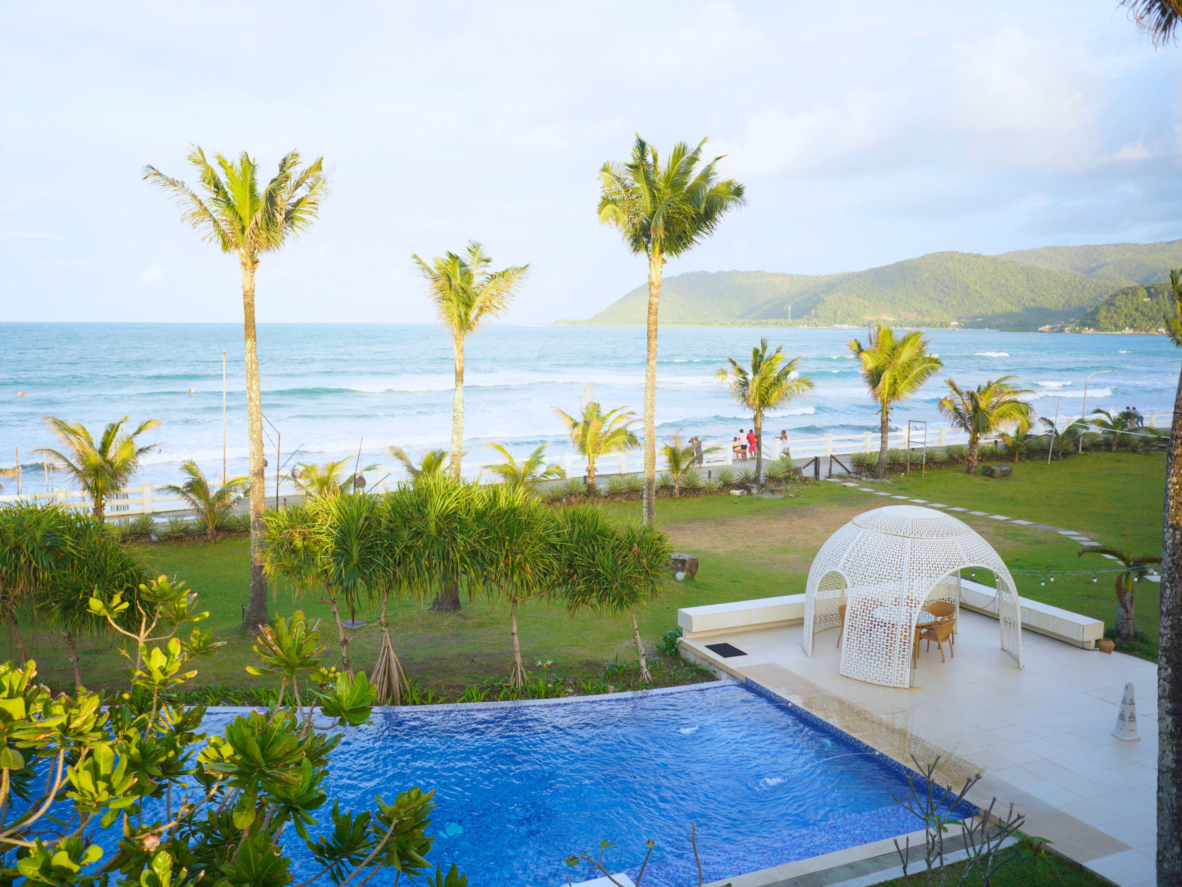Una piscina con vista al océano, rodeada de palmeras y un pabellón blanco.