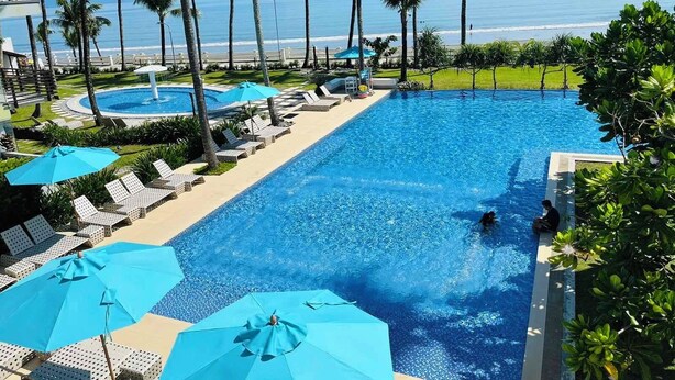 A swimming pool with lounge chairs and umbrellas, surrounded by palm trees and a view of the ocean.