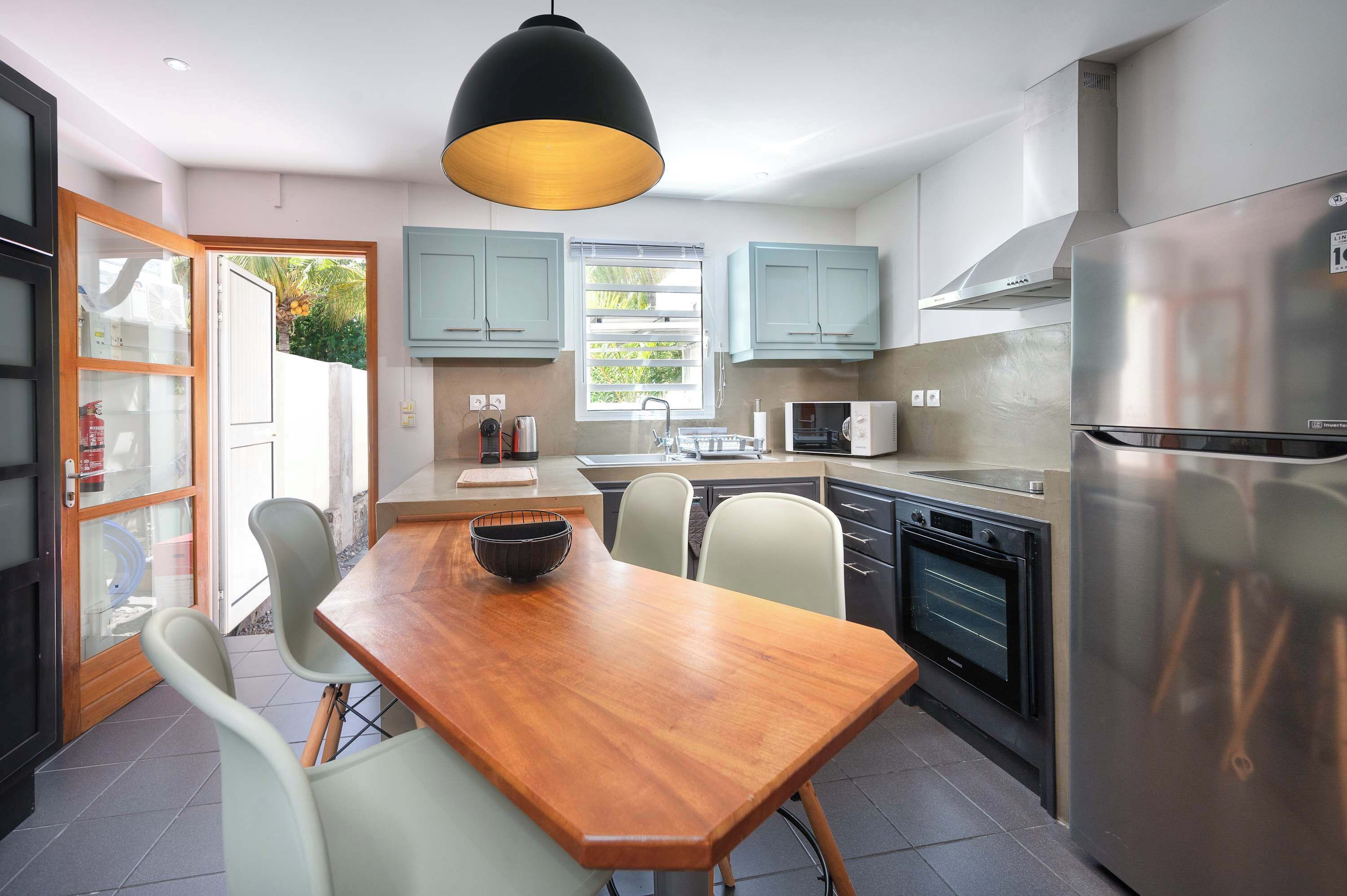 Een moderne keuken met een houten eettafel, lichtgekleurde stoelen, een zwarte hanglamp en roestvrijstalen apparatuur.
