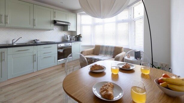 A modern kitchen with light green cabinets, a wooden dining table with plates of food and drinks, and a large window with blinds.