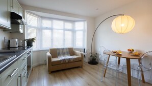 A modern kitchen with a beige sofa, a dining table with chairs, and a large window with blinds.