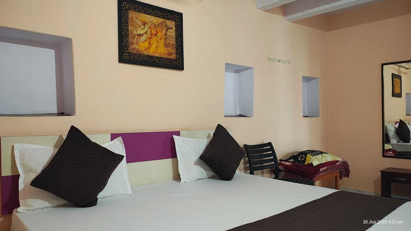 Habitación de hotel con cama, almohadas, una silla, una mesita, un espejo y un cuadro colgado en la pared.