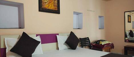 Habitación de hotel con cama, almohadas, una silla, una mesita, un espejo y un cuadro colgado en la pared.