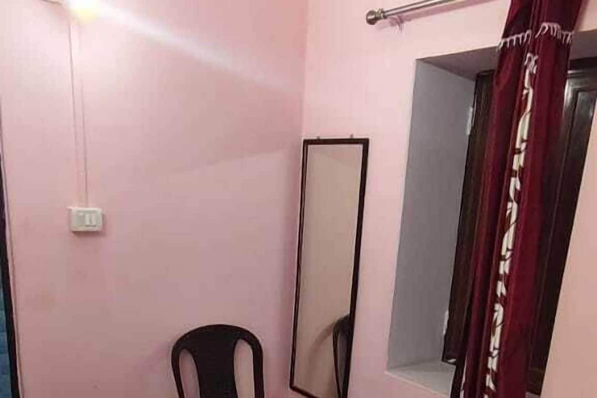 Une petite pièce comprenant un lit, une chaise, un miroir et une fenêtre avec des rideaux.