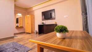 Une table en bois avec un pot de fleurs, un téléviseur fixé au mur et un comptoir d’accueil dans une pièce au plancher en bois et aux murs de couleur claire.