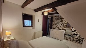 Ein Schlafzimmer mit einem Bett, einem Nachttisch mit Lampe, einer Kommode, einem Fenster und einer Steinwand.