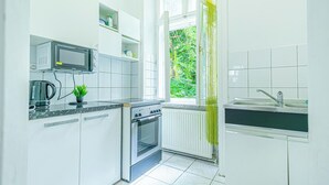A compact kitchen with white cabinets, a built-in microwave, a stove, and a window with a view of greenery. - Simple 2BR Apt near Belvedere Palace (Vienna)