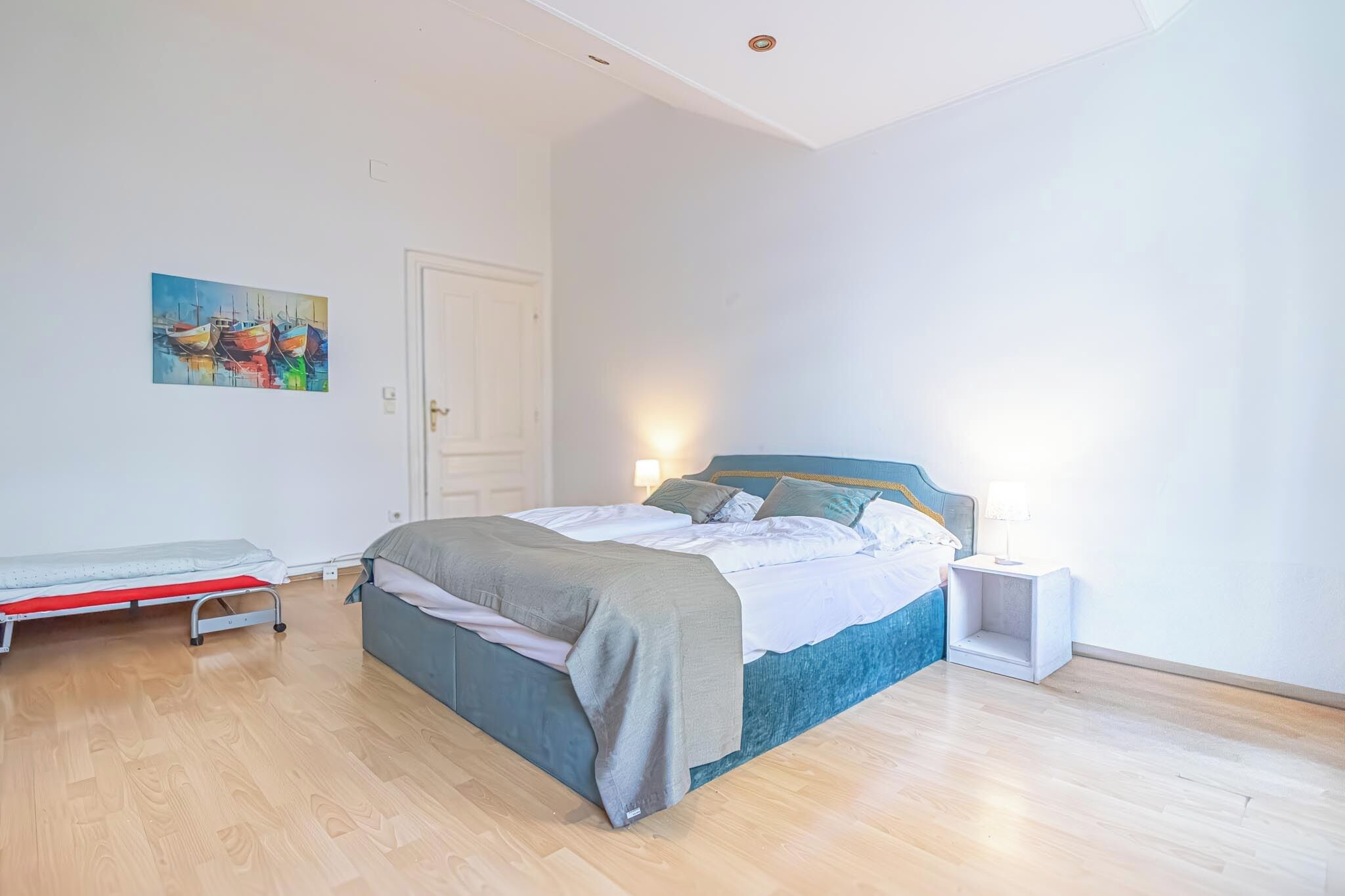 A bedroom with a bed, a bedside table, a nightstand, a wall-mounted shelf, and a door.