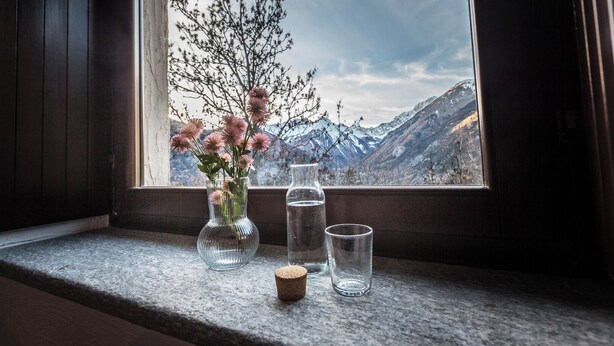 A window with a view of mountains, a vase with flowers, and two glasses on a window sill. - Maison du Chateau (La Salle)