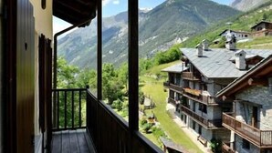 A wooden balcony with a view of a mountain landscape and a building with multiple balconies. - Maison du Chateau (La Salle)