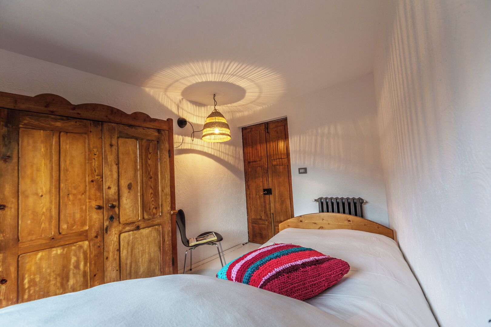 A bedroom with a wooden wardrobe, a bed with a colorful pillow, a hanging lamp, and a radiator.