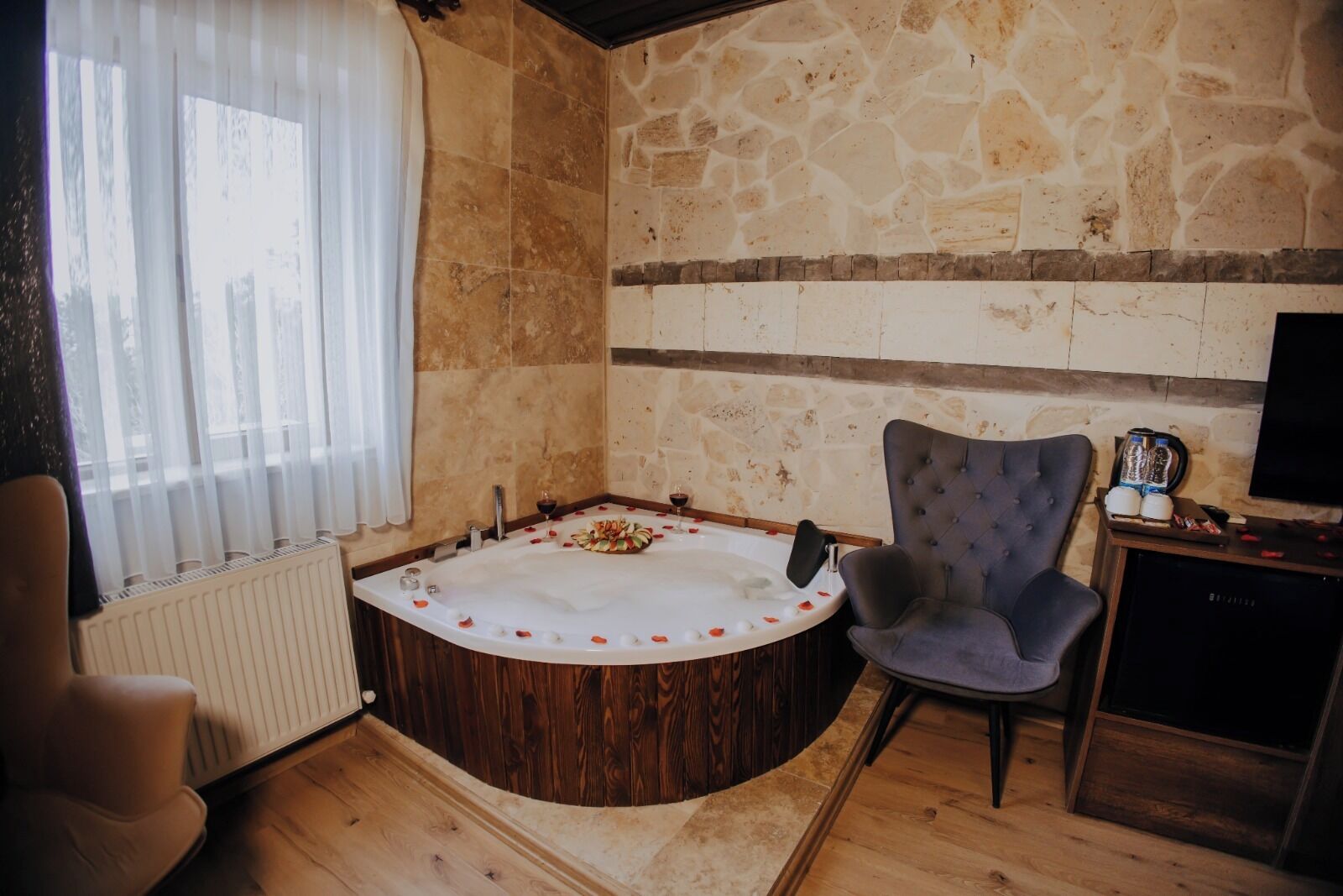 A room with a stone wall, a wooden floor, a round bathtub with rose petals, a grey upholstered chair, and a television on a wooden stand.