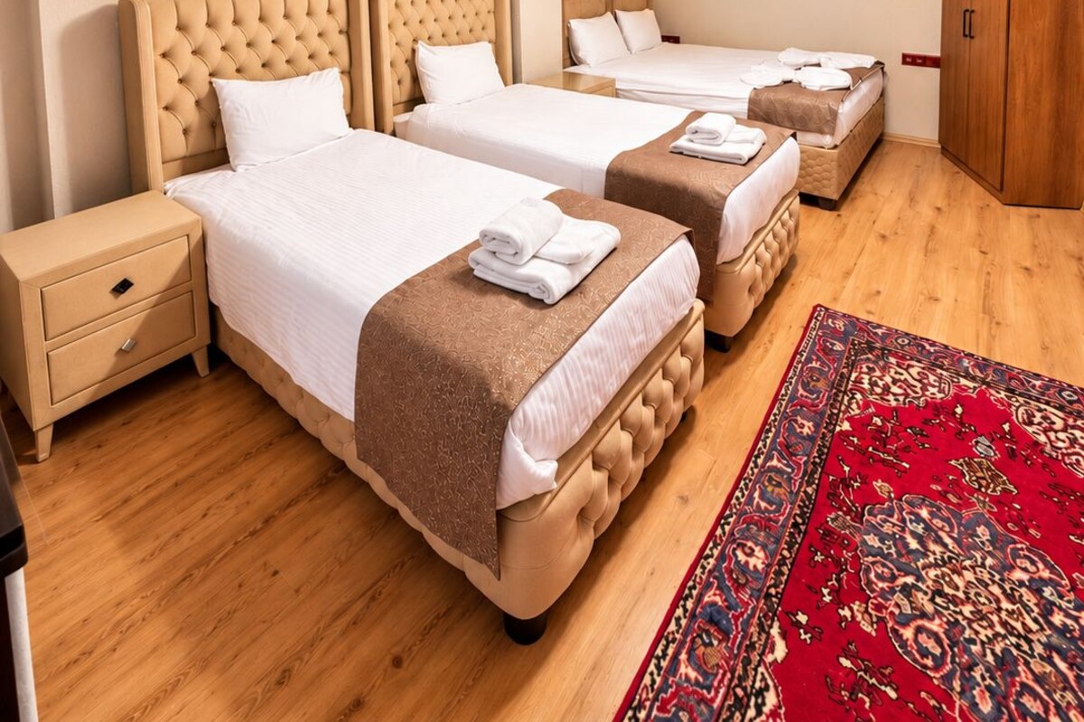 A hotel room with two beds, a wooden ceiling, a chandelier, a wardrobe, and a red patterned rug.
