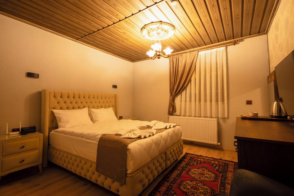 A bedroom with a tufted headboard bed, a bedside table, a radiator, and a wooden ceiling.