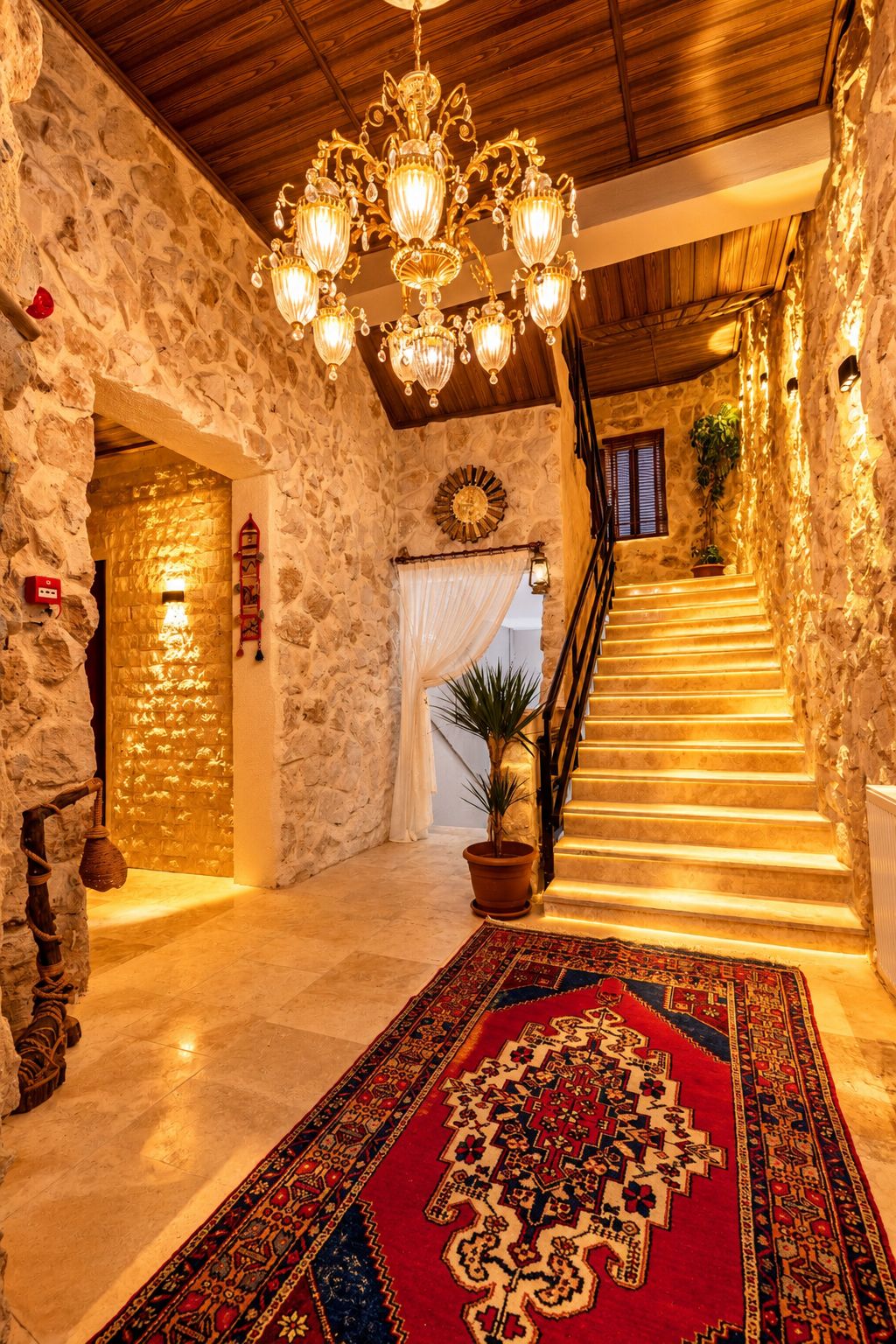 A stone-walled interior with a chandelier, a patterned rug, and a staircase.