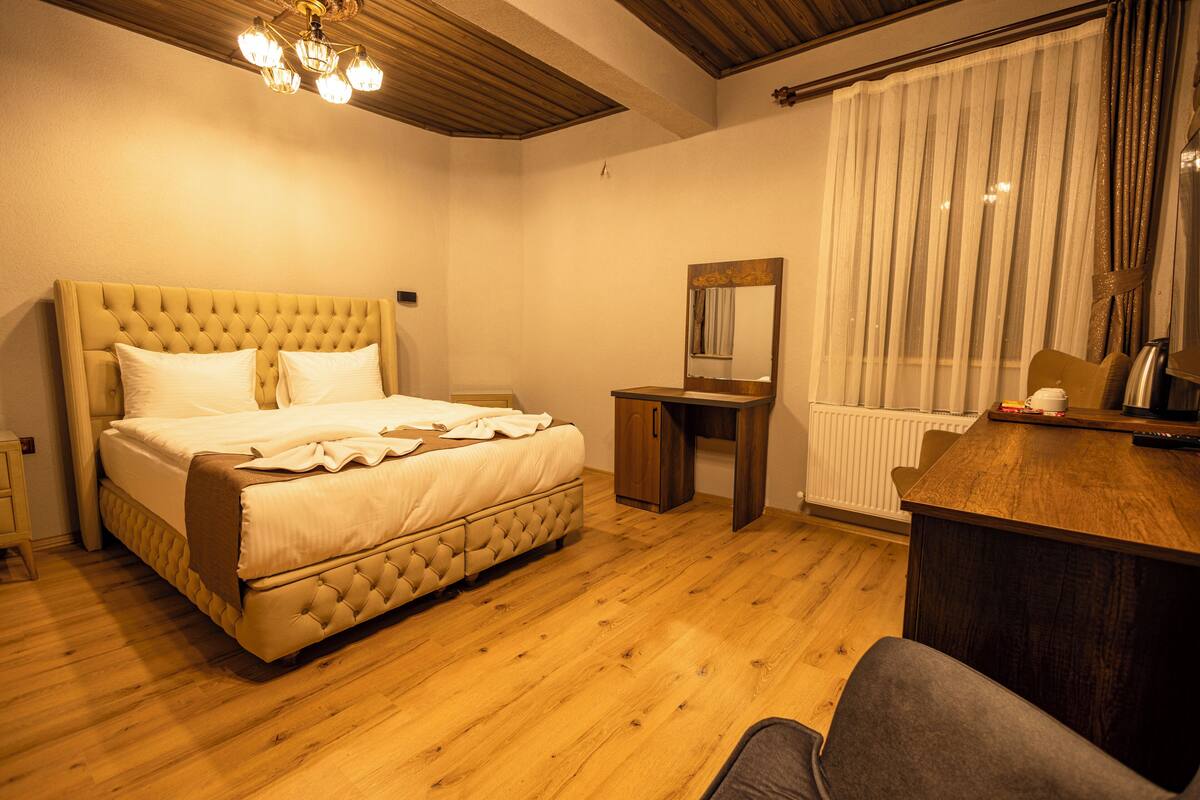A bedroom with a large bed, a wooden desk, a chair, a mirror, and a window with curtains.