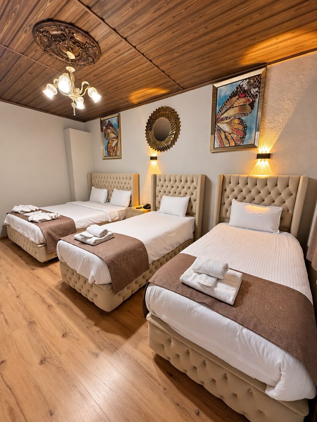 Habitación de hotel con tres camas, suelo de madera, candelabro y cuadros en la pared.